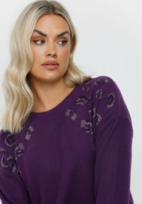 Violetter Pullover mit lockerer Passform, verziert mit einem Leopardenmuster in metallischen Akzenten auf den Schultern. Weicher Stoff.