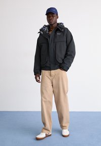 Casaco preto isolado com capuz, forro estampado e dois bolsos frontais. Calças largas bejes e sapatilhas brancas com detalhes castanhos.