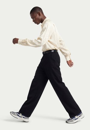 Uomo che cammina indossando una camicia crema a maniche lunghe, pantaloni neri con cintura e scarpe da ginnastica blu navy e bianche su uno sfondo chiaro uniforme.