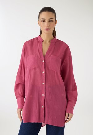 Donna con capelli castani raccolti indietro che indossa una camicia rosa larga, a maniche lunghe e abbottonata, e pantaloni blu scuro su sfondo semplice.