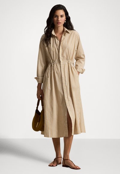 Femme portant une robe chemise mi-longue à rayures verticales beige et blanches, tenant un sac à main marron, chaussée de sandales marron et portant des boucles d'oreilles en perles.