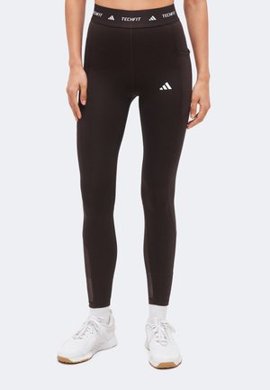 Nő fekete Adidas Techfit leggingset visel oldalzsebbel, fehér zoknit és fehér sportcipőt, egyszínű háttér előtt áll.