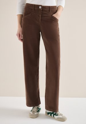 Personne portant un pantalon marron à jambes droites avec une fermeture à boutons, un haut blanc à manches longues et des baskets blanches avec des accents verts.
