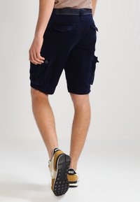 Muž v tmavě námořnických cargo šortkách a béžových teniskách Nike odchází na jednobarevném světlém pozadí.