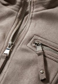 Primo piano di una giacca in suede color taupe con due cerniere metalliche, una principale e una più piccola per la tasca, dotate di tiranti in suede.