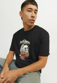 Juodas medvilnės marškinėliai su spalvinga kaukolės grafika, turinčia ragus ir liepsnas, kuriuos lydi tekstas "Jack & Jones" ir "Nuo 1990 metų."