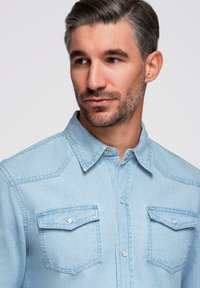Chemise en denim bleu clair avec un col pointu, deux poches poitrine, des fermetures à boutons-pression et une texture subtile du tissu.