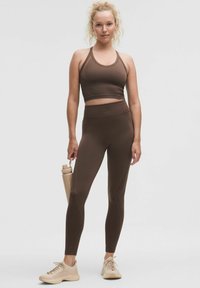 Soutien-gorge de sport marron et legging taille haute en tissu extensible. Une bouteille d'eau beige est tenue. Les chaussures présentent un design en maille.