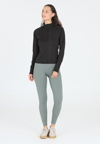 Schwarze Trainingsjacke mit Reißverschluss und langen Ärmeln, kombiniert mit hellgrünen Leggings und beigen Turnschuhen, vor einem schlichten weißen Hintergrund.