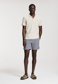 Beige polo póló, kék és fehér csíkos rövidnadrág, zöld szandál. A modell egyenesen áll, karjai lazán lógnak, semleges háttér.