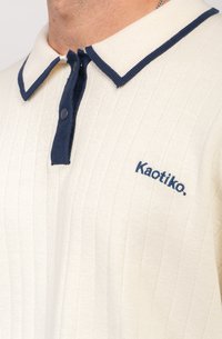 Polo de cuello en color crema con ribetes en azul marino, textura acanalada, dos botones y logo bordado "Kaotiko" en el pecho.