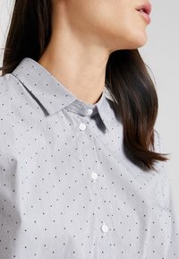 Camicia a bottoni in tessuto a righe blu chiaro e bianche, con piccoli pois neri, colletto e bottoni rotondi bianchi.