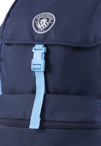 Zaino blu navy con una tasca anteriore con cerniera, dettagli blu chiaro e logo del Manchester City. Realizzato in tessuto resistente con un design strutturato.
