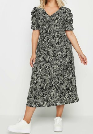 Kobieta ubrana w czarno-białą kwiatową sukienkę midi z bufiastymi krótkimi rękawami i białe trampki, stojąca na jednolitym tle.