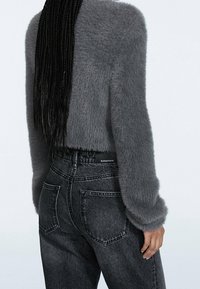 Persona con el pelo largo trenzado, vistiendo un suéter gris borroso y jeans de mezclilla negros de cintura alta, visto desde atrás.