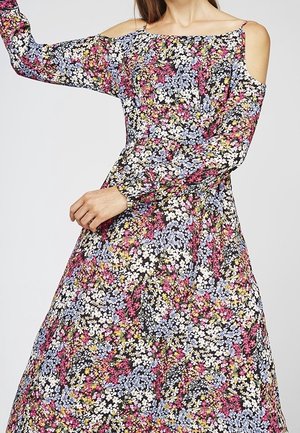 Frau trägt ein langes Blumenmuster-Kleid mit Cutouts an den Schultern und langen Ärmeln, verziert mit kleinen weißen, rosa und blauen Blumen.