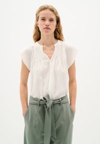 Blusa bianca a maniche corte con scollo a V e dettaglio plissettato sul davanti, abbinata a pantaloni verdi a vita alta con cintura in tessuto e tasche.