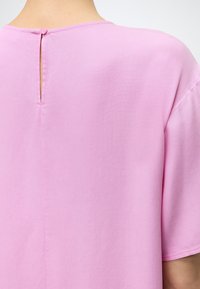 Οπίσθια όψη ατόμου που φοράει ανοιχτό ροζ υφασμάτινη κοντομάνικη μπλούζα με μικρό κουμπί στο άνοιγμα του λαιμού.