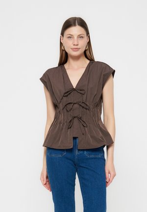 Γυναίκα που φοράει καφέ αμάνικη μπλούζα με V-λαιμόκοψη και τρεις κορδέλες μπροστά, μαζί με μπλε τζιν, στέκεται μπροστά σε απλό ανοιχτό υπόβαθρο.