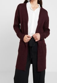 Cárdigan de punto burdeos con abertura frontal, mangas largas y dos bolsillos frontales. Textura suave y corte holgado, llevado sobre una camisa blanca.