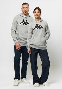 Grijze hoodies met een groot zwart grafisch logo op de voorkant, gecombineerd met donkere spijkerbroeken en witte sneakers. Regular fit, casual stijl.