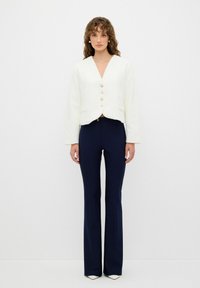Blazer blanco texturizado con escote en V y cuatro botones dorados, combinado con pantalones ajustados de campana azul marino y tacones blancos puntiagudos.