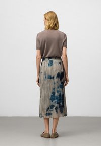 Vrouw met schouderlang blond haar die een bruin kortmouwtopje en een geplooide midi-skirt met een blauw tie-dye patroon draagt, staande met haar rug naar de kijker.