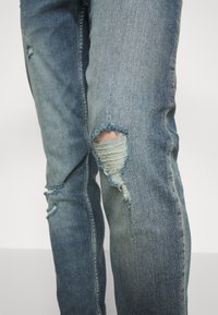 Los jeans de mezclilla oscura presentan desgastes prominentes en las rodillas, una textura desgastada y un corte ajustado. La tela muestra un ligero desvanecimiento y deshilachado.