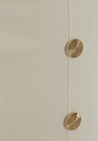 Tela beige con un patrón de tejido diagonal, que presenta un botón dorado prominente con un diseño radial texturizado.