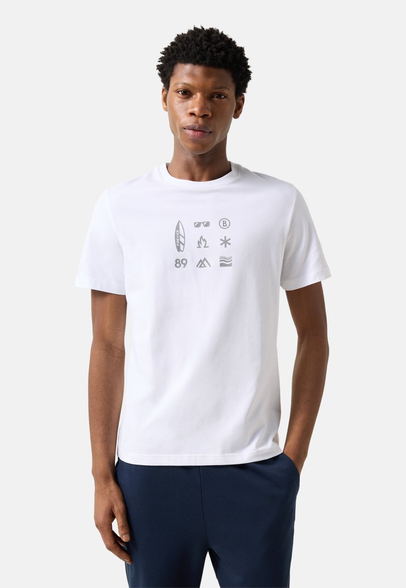 Jeune homme portant un t-shirt blanc avec des icônes grises de plage et d'extérieur et un pantalon bleu marine, debout devant un fond clair uni.