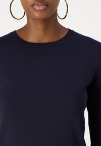 Ναυτικό μπλε πλεκτό πουλόβερ με στρογγυλή λαιμόκοψη, με απαλό σχέδιο και μακριά μανίκια, συνδυασμένο με μεγάλα χρυσά κρίκους σκουλαρίκια.