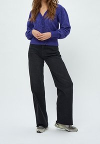Person wearing a purple buttoned cardigan, black wide-leg jeans, and gray sneakers, standing with hands clasped in front.