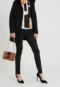 Personne portant un manteau noir, un chemisier à motifs, un pantalon skinny noir, des talons hauts noirs, tenant un sac à main orange imprimé serpent avec une bandoulière chaîne.