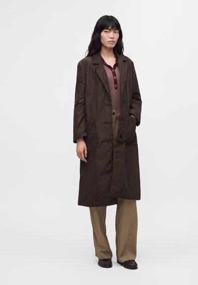 Femme portant un long manteau marron foncé boutonné par-dessus un haut bordeaux avec un pantalon large beige et des chaussures foncées, debout devant un fond uni.