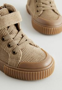 Baskets montantes en velours côtelé beige avec une semelle en caoutchouc, un laçage et une sangle à boucle. Tissu texturé et couture accentuée visibles.