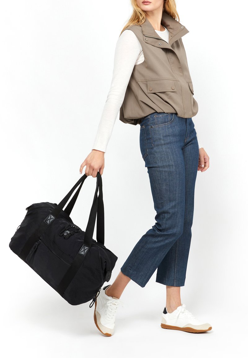 Borsa a duffel nera con due manici superiori, cerniere e dettagli in tessuto. Il modello indossa un gilet beige e jeans blu abbinati a sneakers bianche.