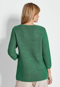 Groene gebreide pullover met een geribbelde structuur, 3/4 mouwen en een ronde halslijn. De lengte reikt voorbij de heupen en heeft een iets losse pasvorm.