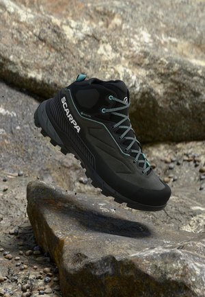 Bota de senderismo Scarpa negra y verde azulado flotando sobre terreno rocoso con cordones visibles y suela resistente para uso al aire libre.