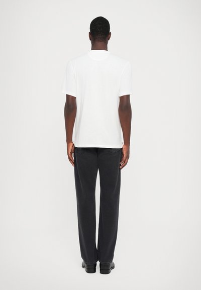T-shirt bianco in cotone con maniche corte e scollo rotondo, abbinato a pantaloni larghi neri e scarpe nere. Lo sfondo è semplice.