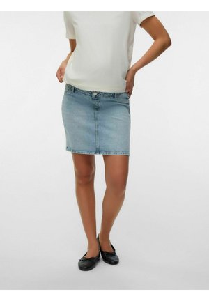 Femme portant un haut blanc à manches courtes, une jupe en jean bleu clair et des ballerines noires, debout les mains sur les hanches.