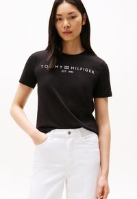 Fekete rövid ujjú póló fehér "TOMMY HILFIGER EST. 1985" felirattal. Fehér farmernadrággal párosítva, laza, kényelmes szabással.
