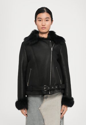 Vrouw in een leren jack met zwarte bontkraag, voorzien van ritssluitingen op de zakken en manchetten, gecombineerd met een asymmetrische spijkerrok in twee kleuren.
