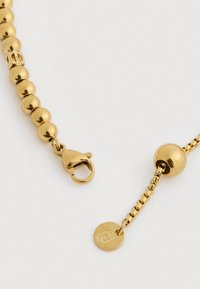 Χρυσός κρεμαστός περιλαίμιος με αλυσίδα και κούμπωμα τύπου αστακού, καθώς και μικρή στρογγυλή ετικέτα με εγχαραγμένο γεωμετρικό λογότυπο σε λευκό φόντο.