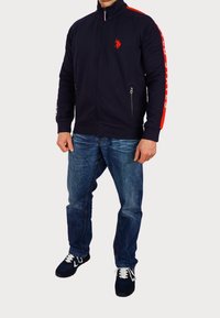 Marinblå zip-up jacka med röda accenter, med en broderad logotyp, sidofickor och ribbade manschetter, i kombination med blå jeans och svarta sneakers.