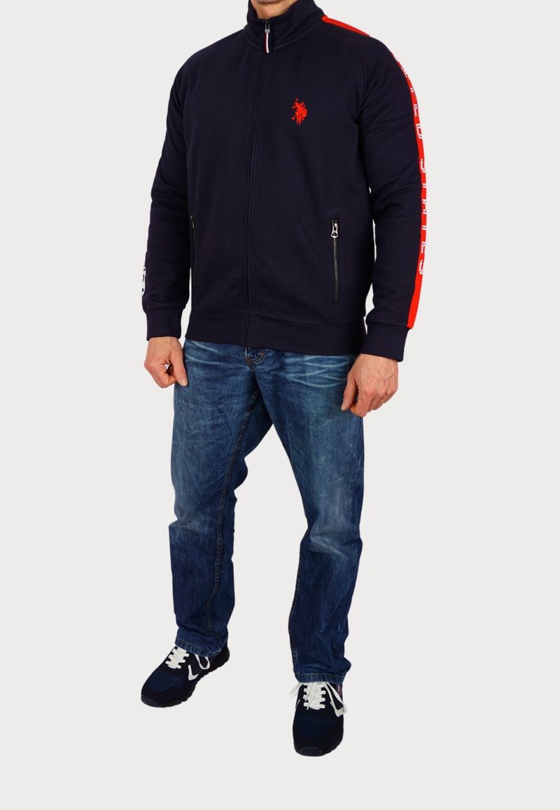 Marinblå zip-up jacka med röda accenter, med en broderad logotyp, sidofickor och ribbade manschetter, i kombination med blå jeans och svarta sneakers.