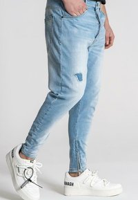 Ljusblå denimjeans med slim fit, en sliten reva på låret och dragkedjor vid vristerna. Matchas med vita sneakers.