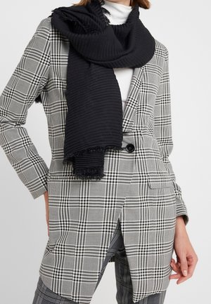 Kvinde iført sort ribstrikket tørklæde over hvid rullekrave og lang sort-hvid ternet blazer med matchende ternede bukser.