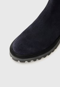 Bota de ante azul marino hasta la rodilla con punta redonda, superficie texturizada y suela de goma negra gruesa con patrones de relieve profundos.
