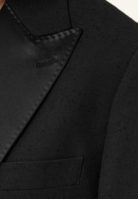Blazer nero con risvolto in satin e tessuto strutturato, che mette in mostra dettagli del foro per il bottone e della tasca sul petto.