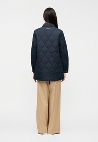 Μπλε ναυτικό καπιτονέ παλτό με ραμμένο γιακά, μανσέτες με κουμπιά και διακριτικό λογότυπο στην πλάτη, συνδυασμένο με φαρδιά μπεζ παντελόνια.